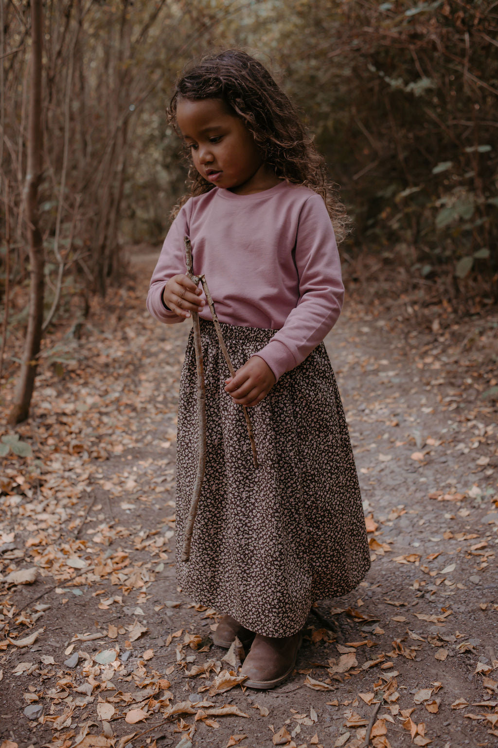 Child in a pink sweater and leopard print skirt standing in a forest path with autumn leaves.