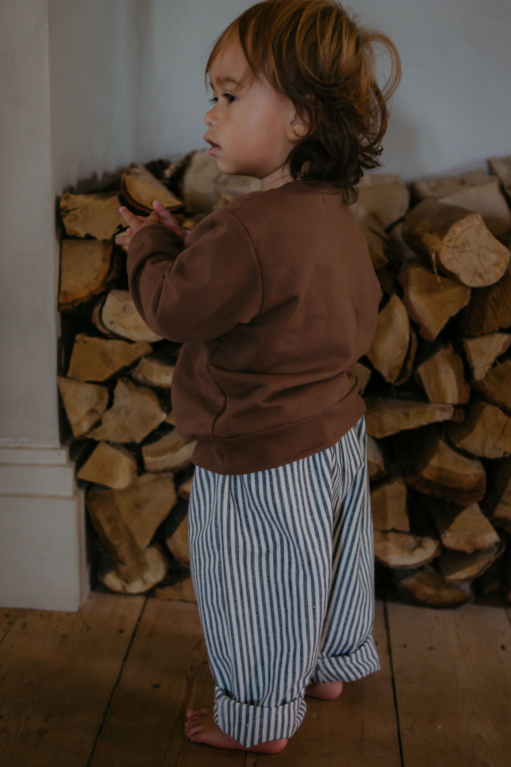 Child in a brown sweater and striped pants standing in front of stacked firewood.