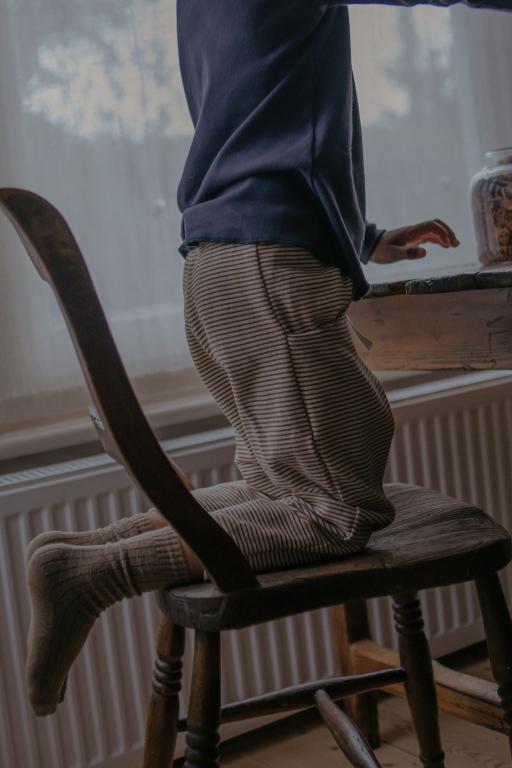 Person standing on a wooden chair in a room with a window and radiator.