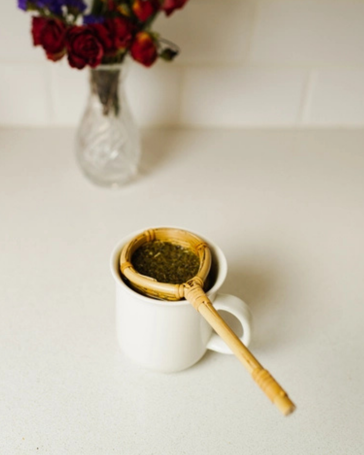 White mug with a bamboo tea infuser and a vase of flowers on a light surface