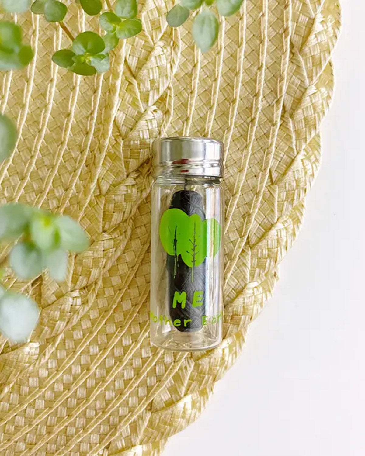 A clear glass container with a label that reads 'Me Mother Earth' and a piece of bamboo charcoal dental floss inside, placed on a woven mat with a yellow background and green leaves in the background.