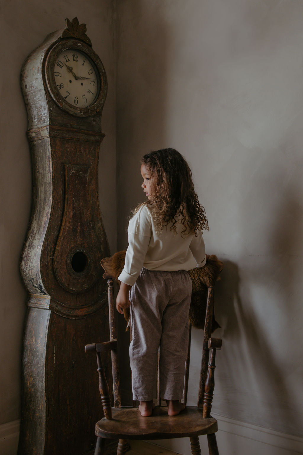 Child standing on a wooden stool next to a large vintage clock against a plain wall.
