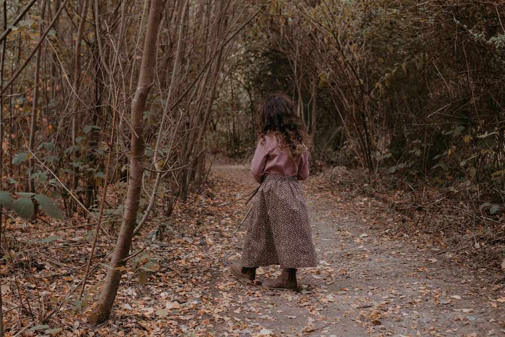 Person walking alone on a path through a forest with bare trees and fallen leaves.