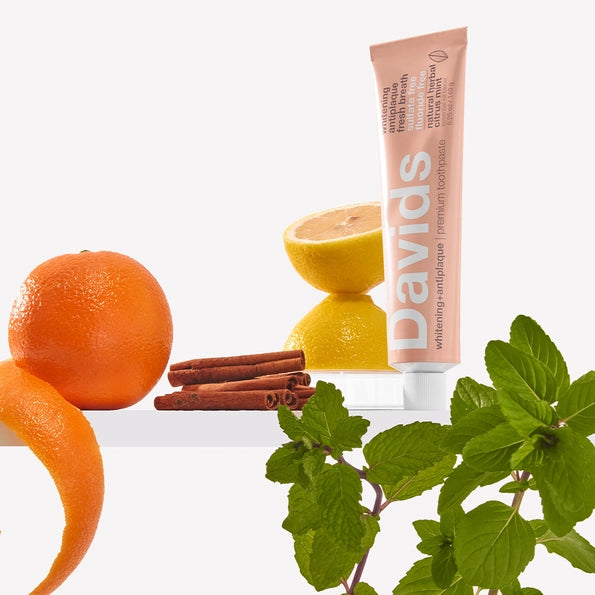 Tube of Davids toothpaste with oranges, a lemon, cinnamon sticks, and mint leaves on a white background